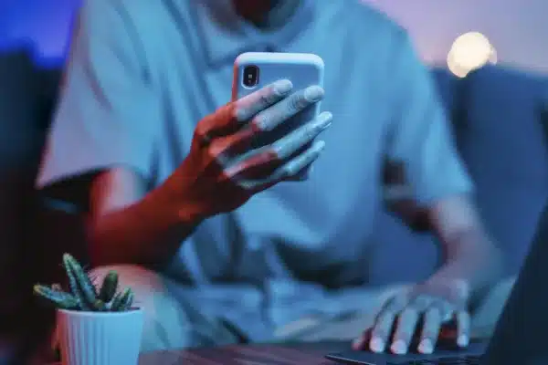 Homem segurando smartphone em um ambiente de trabalho, com laptop e uma pequena planta ao seu lado. Representa o uso de aplicativos de e-commerce no dia a dia dos brasileiros, refletindo a popularidade e a conveniência desses serviços no país.