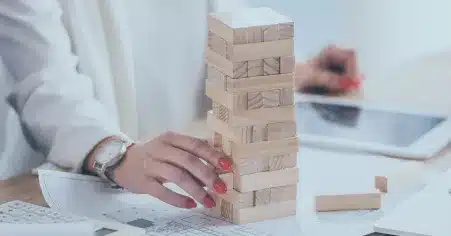 A imagem mostra uma pessoa construindo uma estrutura de blocos de madeira, um por um, em uma mesa. Em segundo plano, há um laptop e papéis, o que sugere um ambiente de trabalho ou de estudo. A pessoa está focada na tarefa, o que indica uma atenção meticulosa e cuidadosa. No contexto do material "Gestão de risco: um guia para aplicar no seu negócio", a imagem pode ser usada para ilustrar a importância de construir uma gestão de risco sólida e estratégica para o negócio, assim como os blocos de madeira precisam ser cuidadosamente colocados para construir uma estrutura estável.