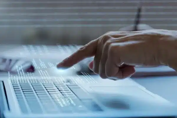 A imagem exibe uma mão apontando para uma telinha de laptop, que exibe uma interface de software com várias opções e menus. A imagem é em tons azuis e parece ter um efeito de luz digital em torno do dedo, o que sugere interação com a tecnologia. No contexto do artigo "Passo a passo da implementação do ERP ONCLICK na sua empresa", a imagem pode representar o processo de seleção e configuração de funções do ERP (Sistema de Planejamento Empresarial). A mão apontando para a tela pode simbolizar a escolha de módulos ou a personalização do ERP para atender às necessidades específicas da empresa. A interface digital reflete a natureza tecnológica e a complexidade do software ERP, que requer uma implementação cuidadosa e uma compreensão das diferentes funcionalidades para ser eficaz. A imagem ilustra a ideia de que a implementação do ERP é um ato intencional e estratégico, envolvendo a seleção de recursos e a configuração do sistema para integrar-se ao fluxo de trabalho da empresa.