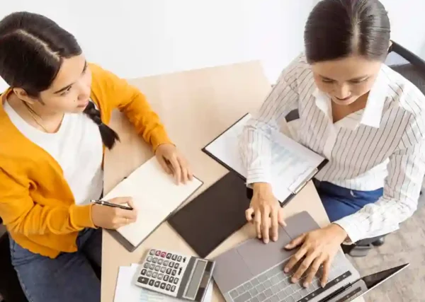 Visão superior de duas profissionais trabalhando juntas em uma mesa de escritório clara. Uma delas, vestindo uma blusa amarela, faz anotações em um caderno, enquanto a outra, vestindo uma camisa listrada, opera um notebook e analisa documentos em uma prancheta. Sobre a mesa, há uma calculadora financeira e papéis. A imagem ilustra a integração contábil e a colaboração entre equipes financeiras, destacando a precisão na gestão fiscal e a automação de processos possibilitada pelo sistema ERP da ONCLICK.
